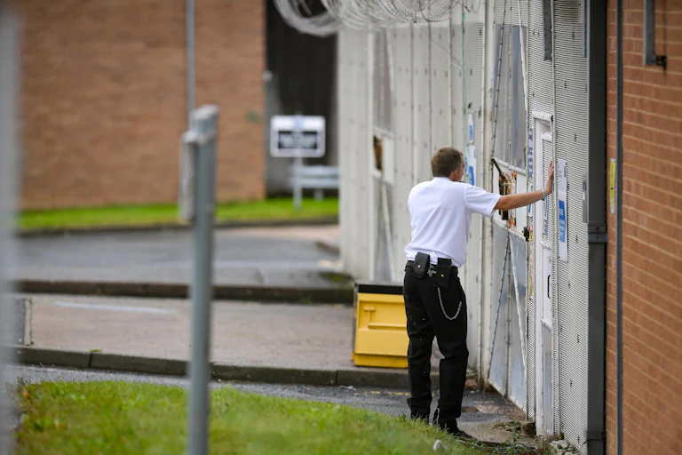 A prison officer leans against a gate outside of a prison