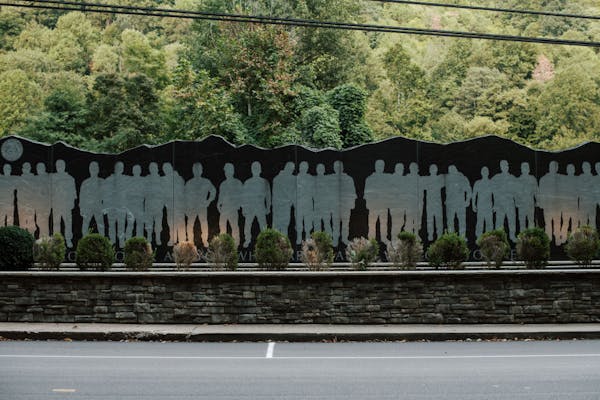 A black wall with paler silhouettes of men standing along it, positioned above a low stone wall next to the road.