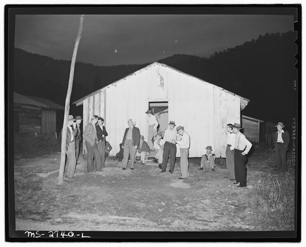 A black and white photo of several men in slacks and shirts standing outside a small white building.