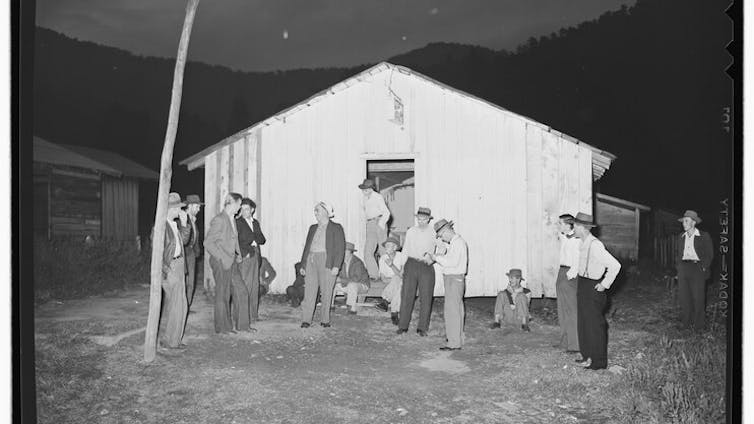 A black and white photo of several men in slacks and shirts standing outside a small white building.