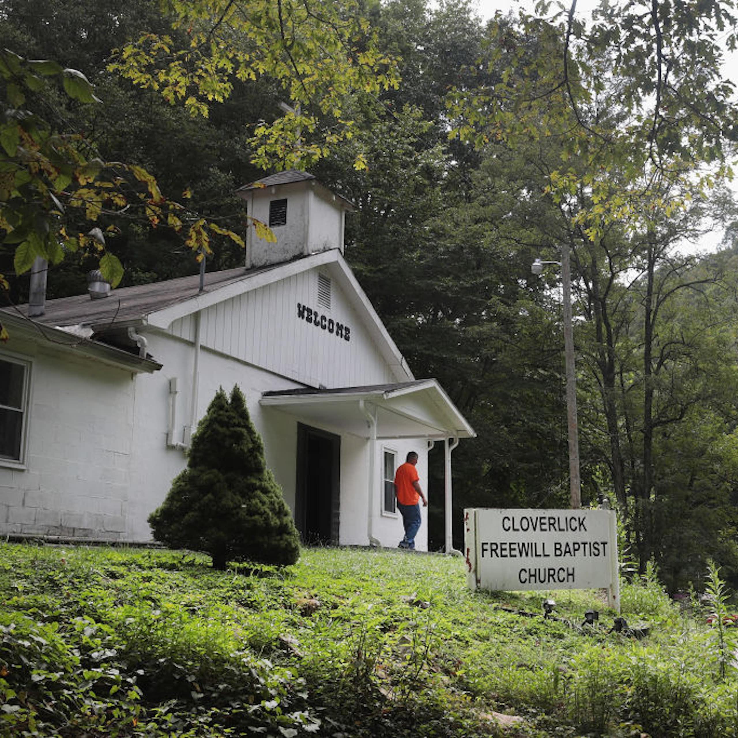 A man in an orange shirt walks away from a small white church in a forested area.