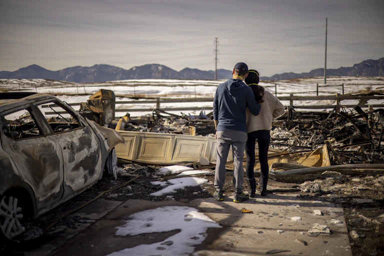 A man and woman stand side-by-side in front of the wreckage of a fire.