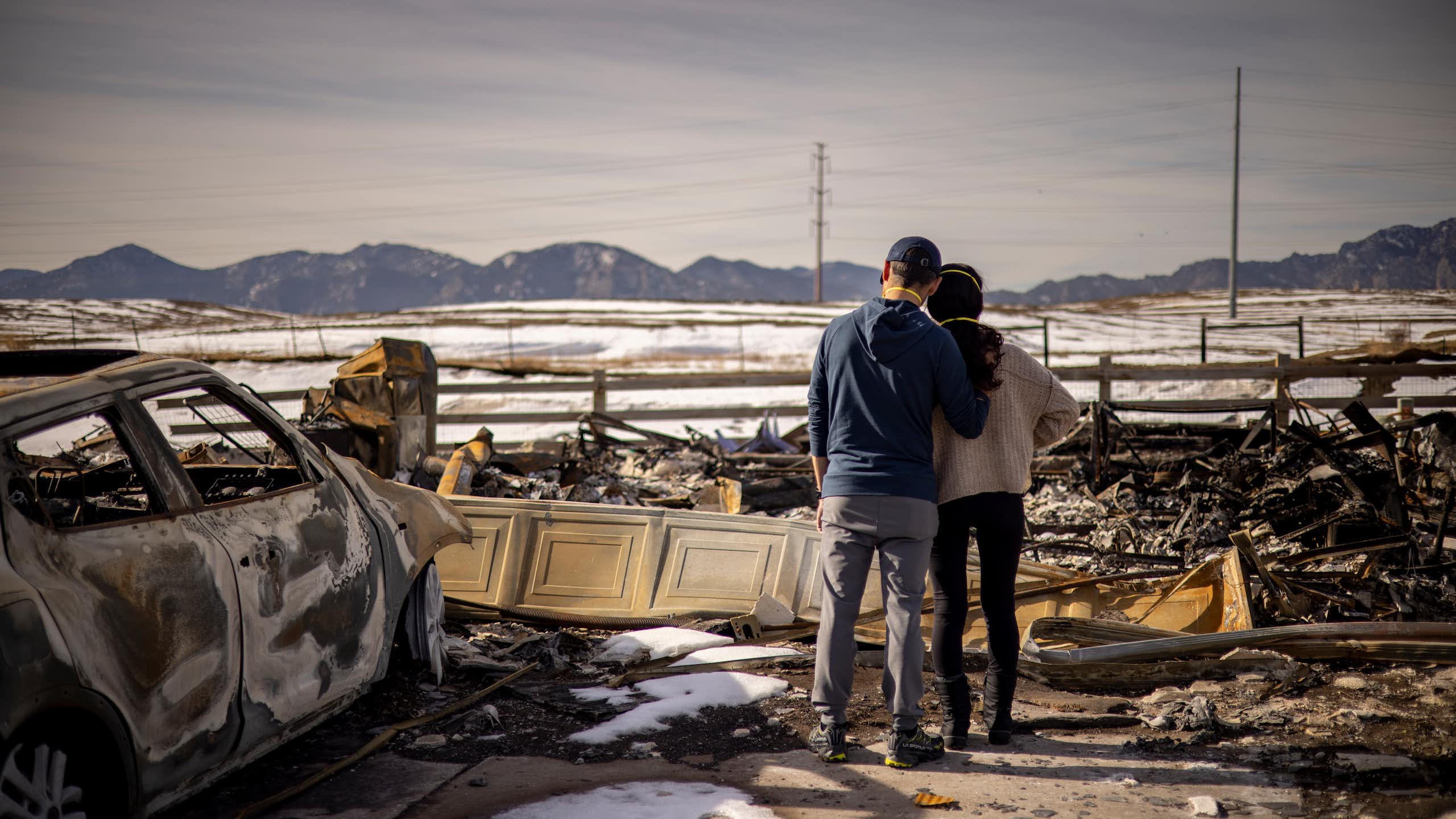 A man and woman stand side-by-side in front of the wreckage of a fire.