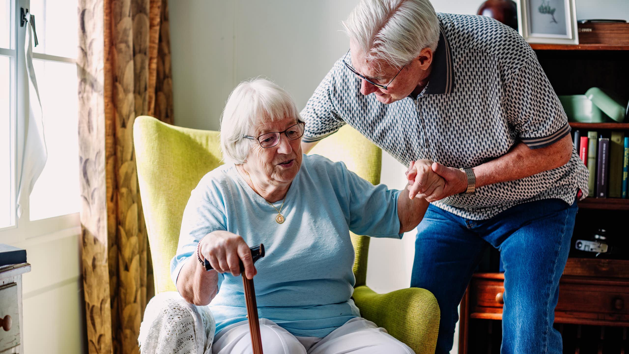 Une dame âgée s’appuie sur une canne pour se lever d’un fauteuil, aidée par un homme âgé.