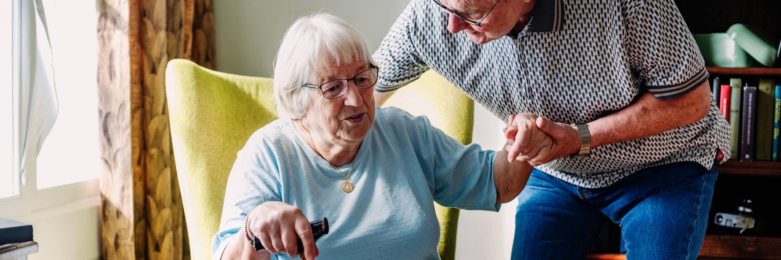 Une dame âgée s’appuie sur une canne pour se lever d’un fauteuil, aidée par un homme âgé.