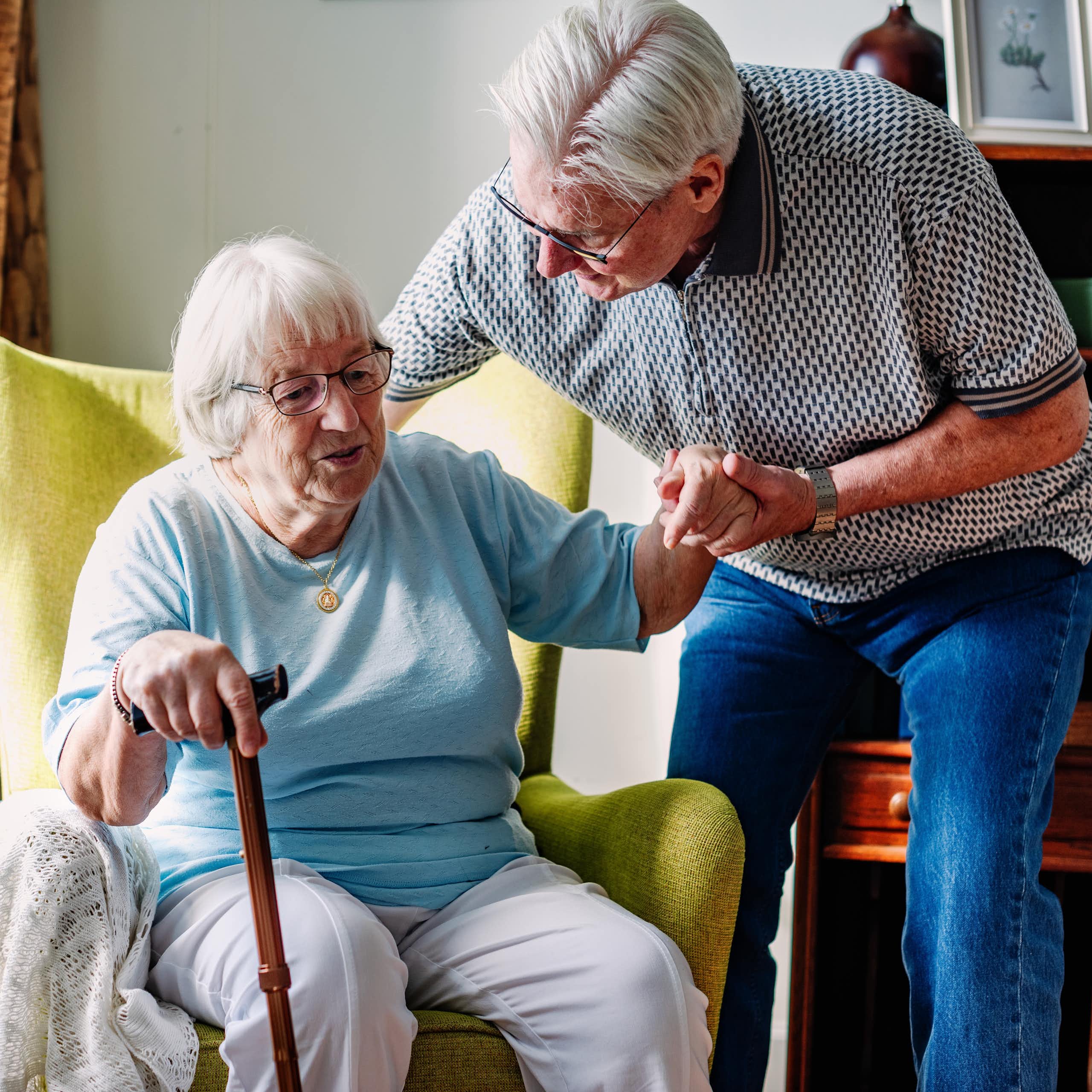 Une dame âgée s’appuie sur une canne pour se lever d’un fauteuil, aidée par un homme âgé.