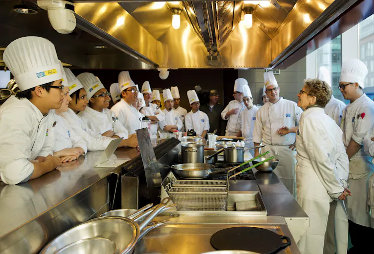 People in chef hats and whites seen around a kitchen with a woman in a suit and glasses.