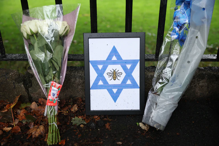 A framed Star of David with a bee, and two bouquets of flowers left up against a fence