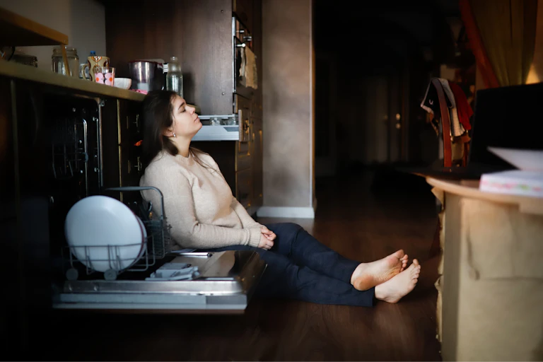 Exhausted woman slumps on kitchen floor near open dishwasher.