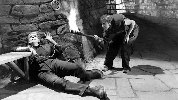 Un hombre deforme yace en el suelo y otro con joroba lo observa iluminándose con una antorcha.