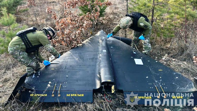 A black drone lies on the ground while two soldiers inspect it