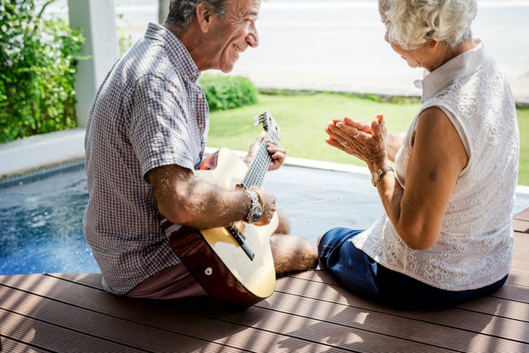 an older man sits playing a guitar as a grey-haired woman smiles and watches