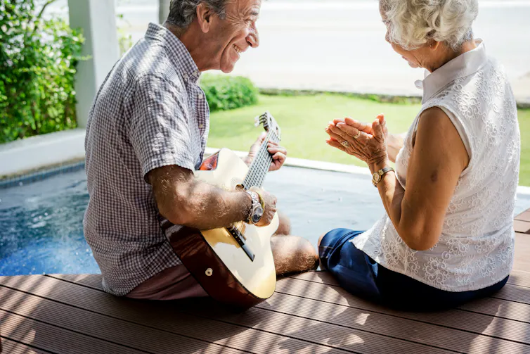an older man sits playing a guitar as a grey-haired woman smiles and watches