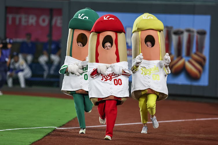 Three people dressed up in hot dog costumes – one green, one red and one yellow – race during a baseball game.