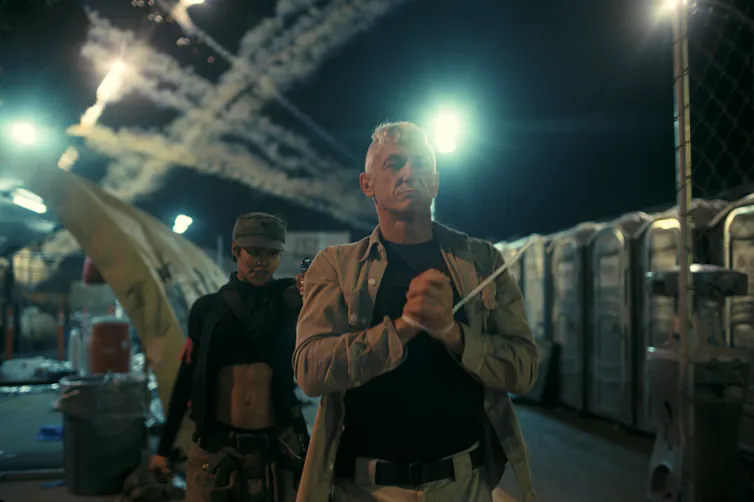 A man with his hands tied walking ahead of a woman with a gun pointed at his head