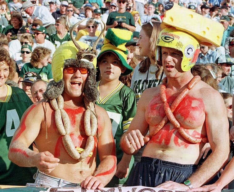 Two shirtless fans, one wearing a cheesehead hat, another wearing a helmet with antlers. Both wear sashes made of sausage links.