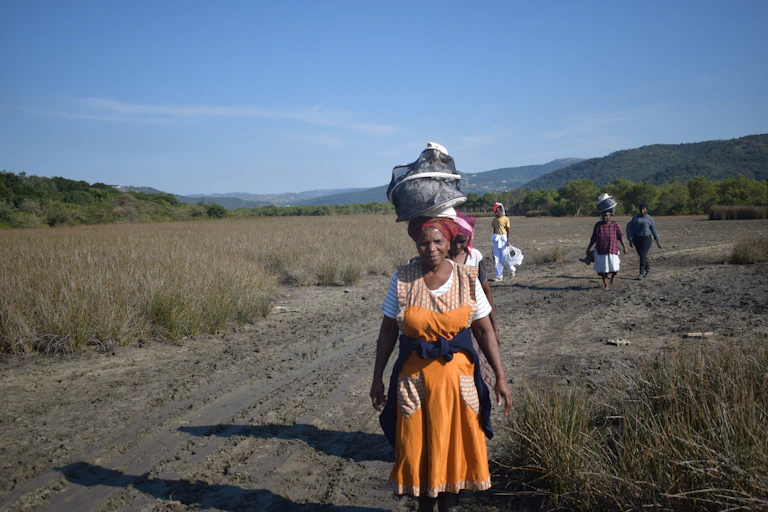 Four women walk along a long dirt road carrying heavy loads on their heads