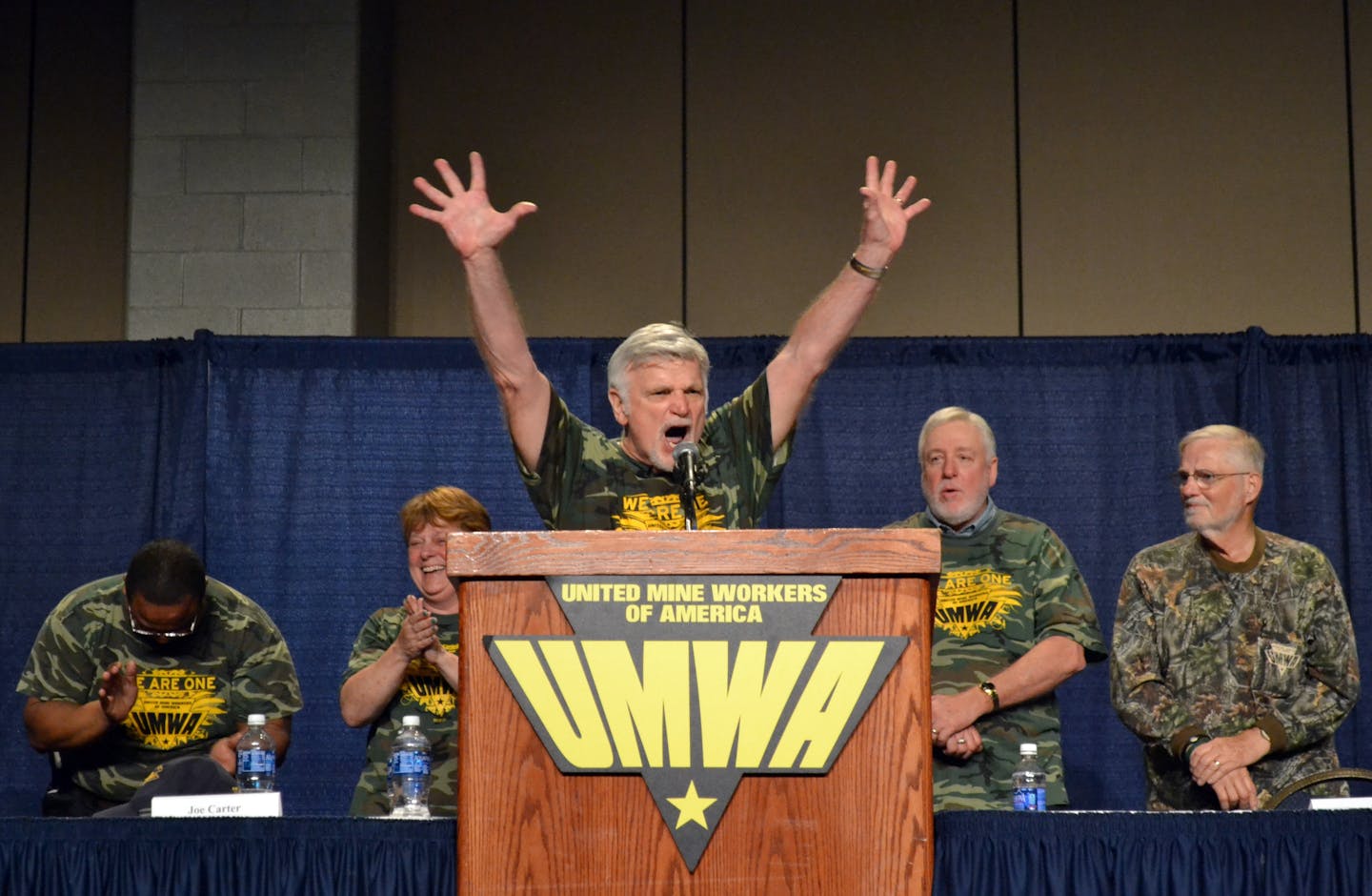 A man with white hair raises his arms as he speaks animatedly at a podium, while he and other men nearby wear camo-colored t-shirts.