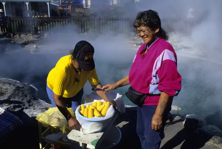 Two people stand near a large container of shucked corn while steam billows from a pool of water behind them.