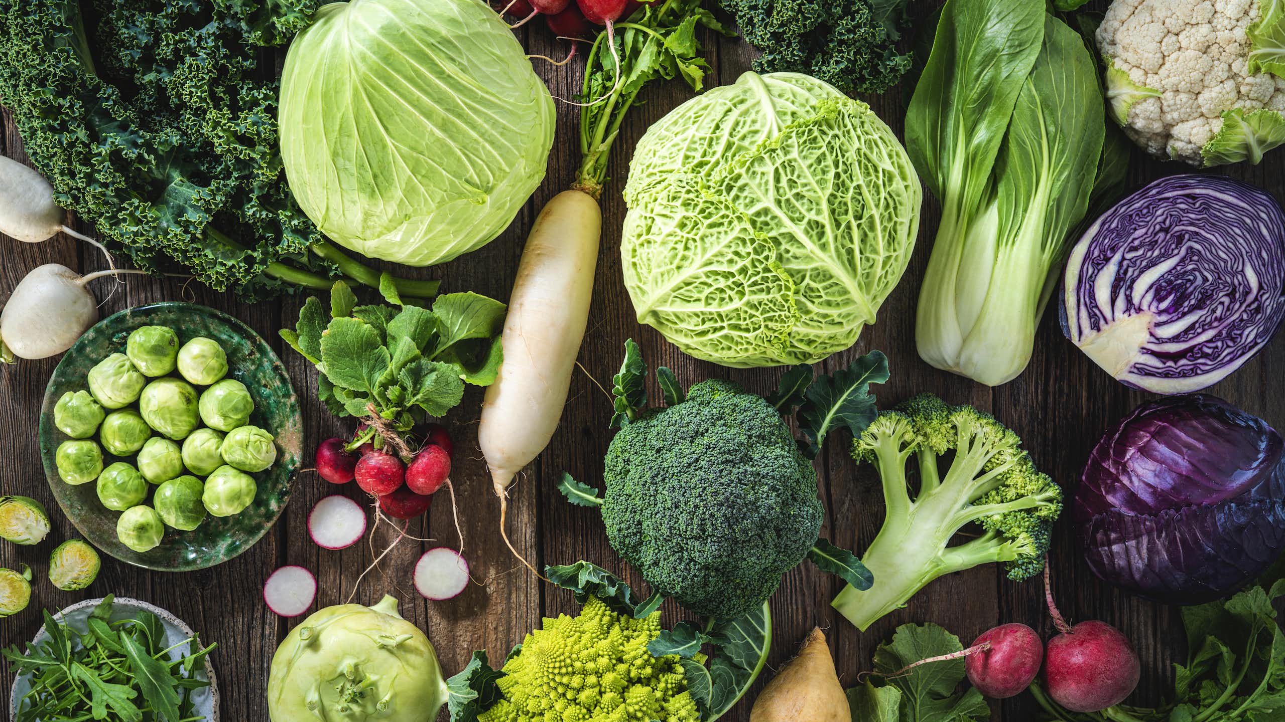 An assortment of vegetables including cabbage, broccoli, cabbage, turnip, kale, romanesco, radish, arugula, kohlrabi and swedes on wood rustic board