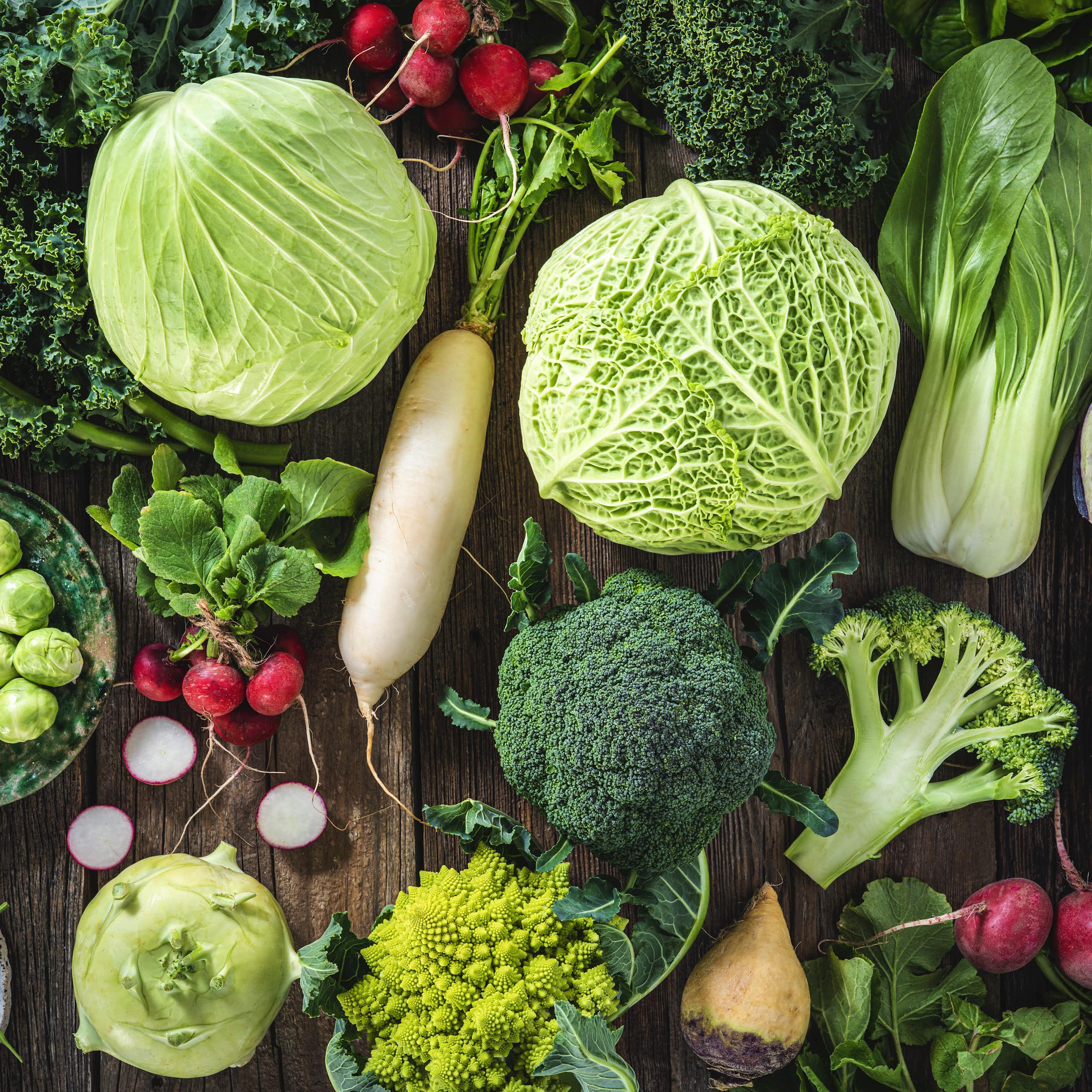 An assortment of vegetables including cabbage, broccoli, cabbage, turnip, kale, romanesco, radish, arugula, kohlrabi and swedes on wood rustic board