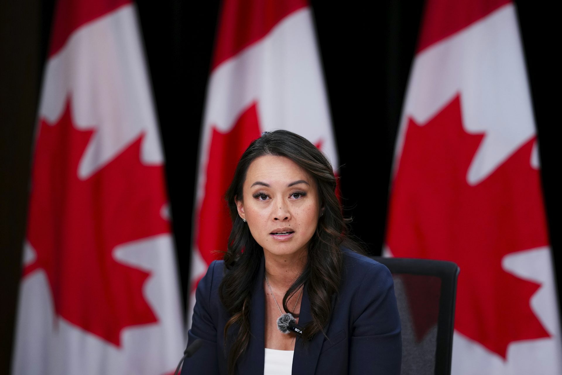 A Filipino woman with long, dark hair in a blazer speaks to someone off-screen while sitting in front of a row of Canadian flags