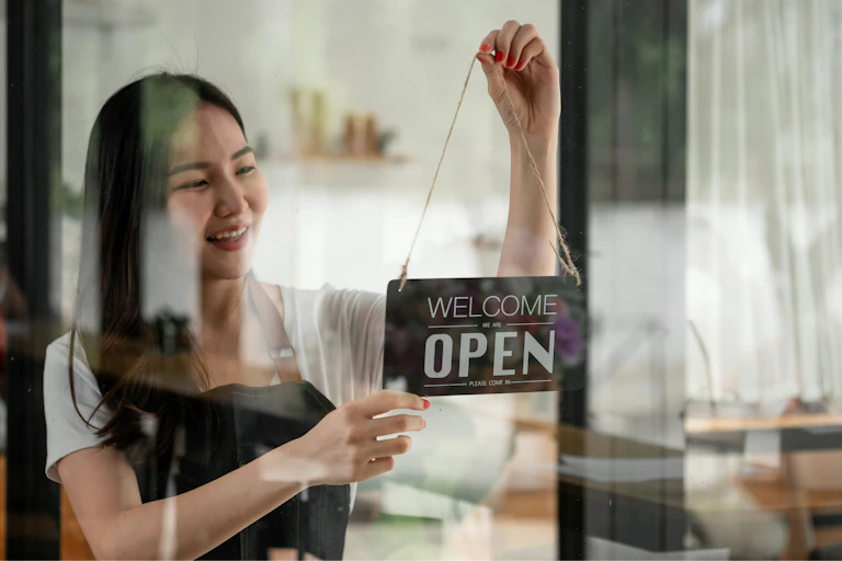 A woman hands a 'Welcome we are open' sign in the doorway of a shop