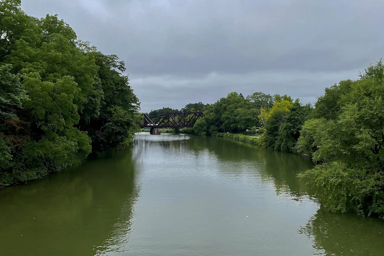 An outdoor scene of a calm river surrounded by banks with leafy trees, and a rust-colored bridge in the distance.