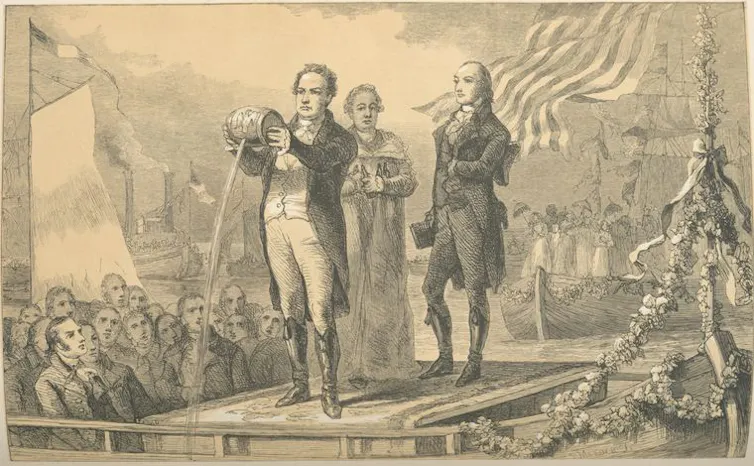 A faded black-and-white illustration of men in formal 18th-century clothing standing in front of a crowd as one pours a stream of water from a small barrel.