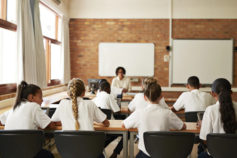 A back view of a classroom. A teacher stands at the front. Students wear white shirts and sit at desks.