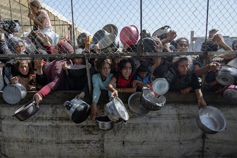 Children holding saucepans clamor for food behind a wire fence in Gaza.