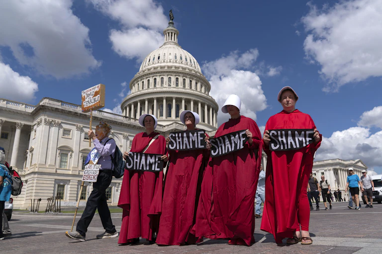Four women in crimson robes walk in front of the Capitol holding signs that read, 'Shame.'