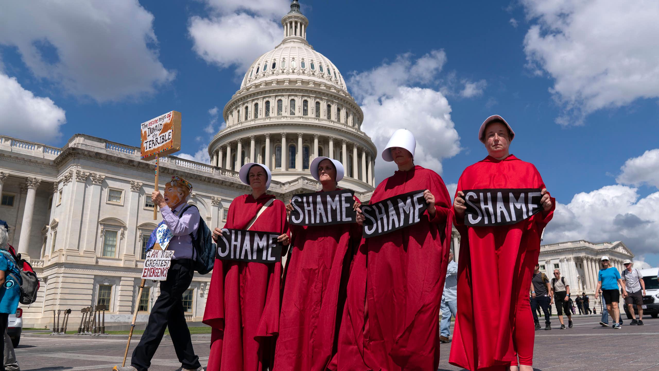 Four women in crimson robes walk in front of the Capitol holding signs that read, 'Shame.'