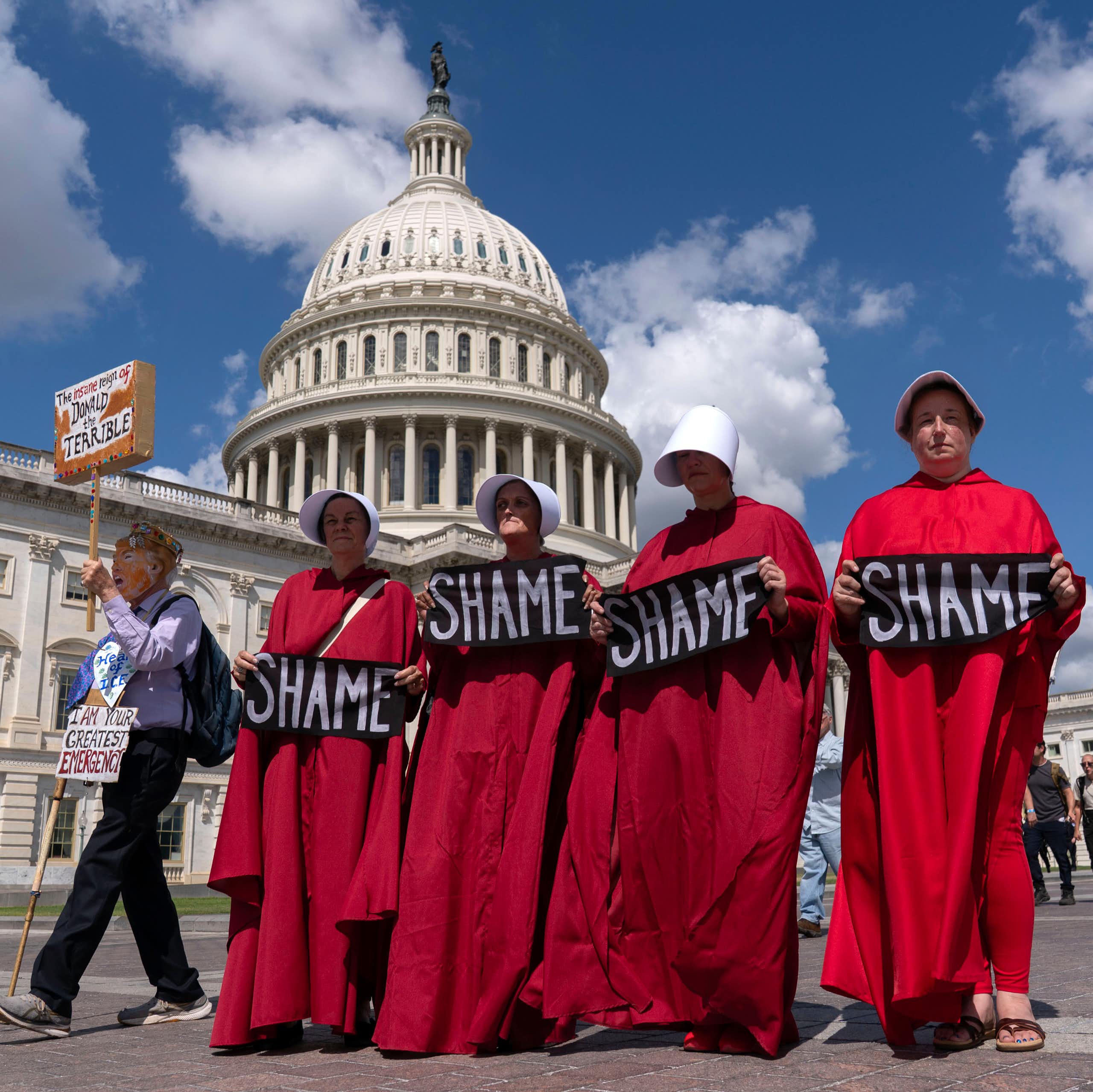 Four women in crimson robes walk in front of the Capitol holding signs that read, 'Shame.'