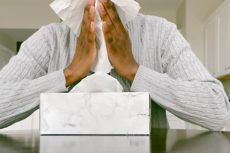 Person sitting at a table with a tissue box, with tissues covering their face.