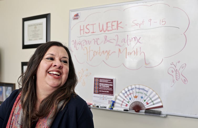 A woman with dark hair smiles and stands in front of a whiteboard that says in marker, 'HSI week, Sept 9-15'