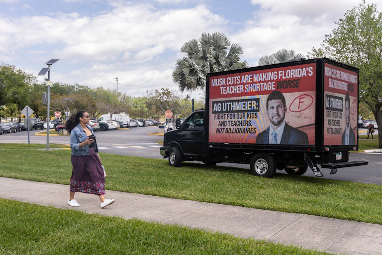 A woman walks past a truck that has a red billboard that says 'Musk cuts are making Florida's teacher shortage worse.'