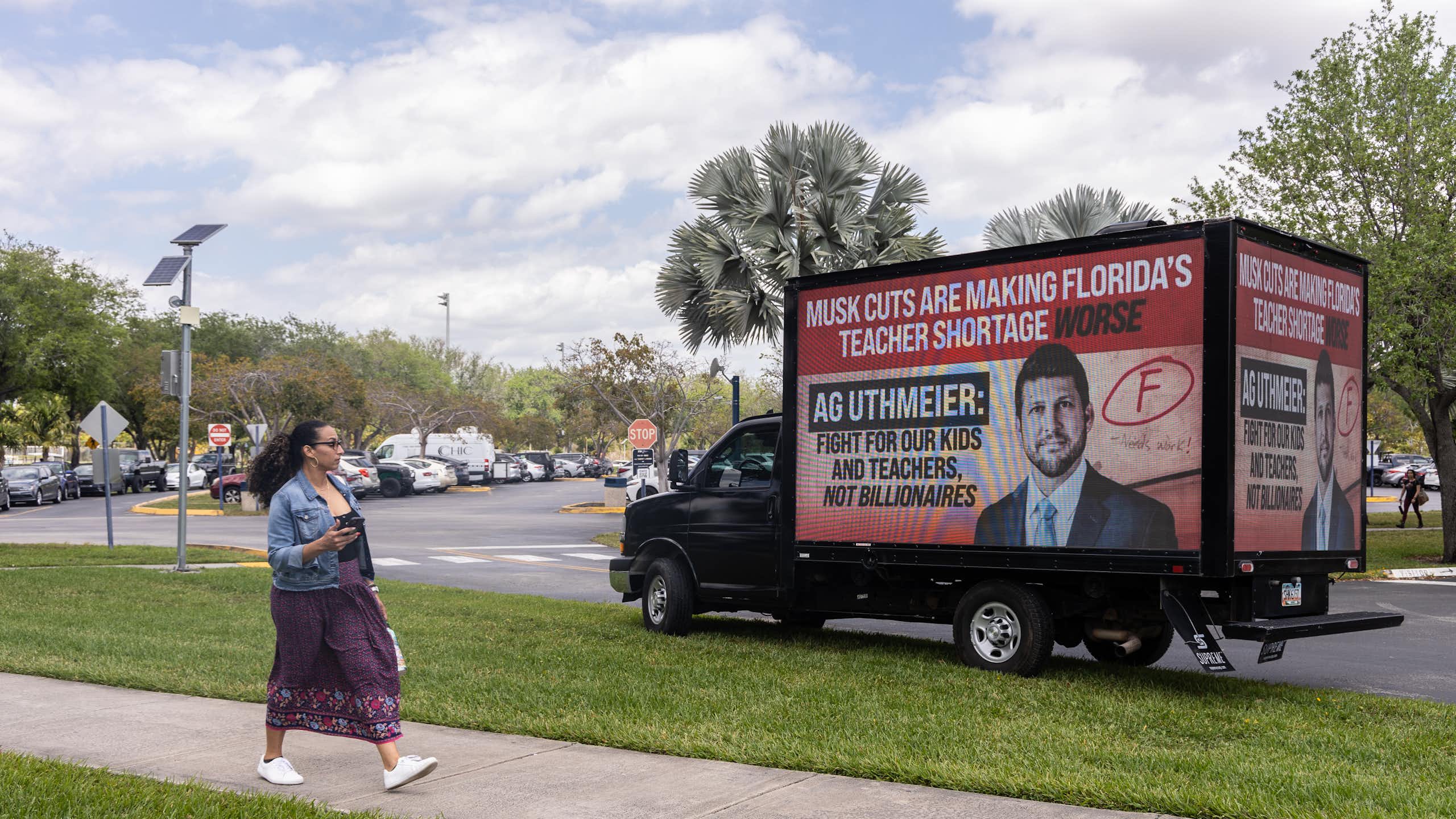 A woman walks past a truck that has a red billboard that says 'Musk cuts are making Florida's teacher shortage worse.'