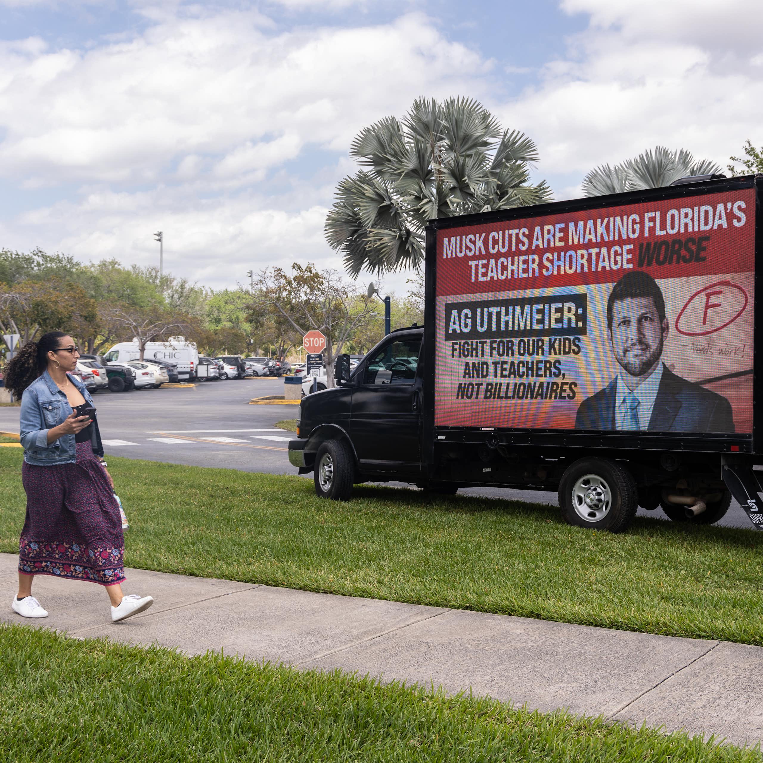 A woman walks past a truck that has a red billboard that says 'Musk cuts are making Florida's teacher shortage worse.'