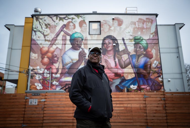 A Black man in sunglasses stands in front of a mural on a city wall featuring Black people.