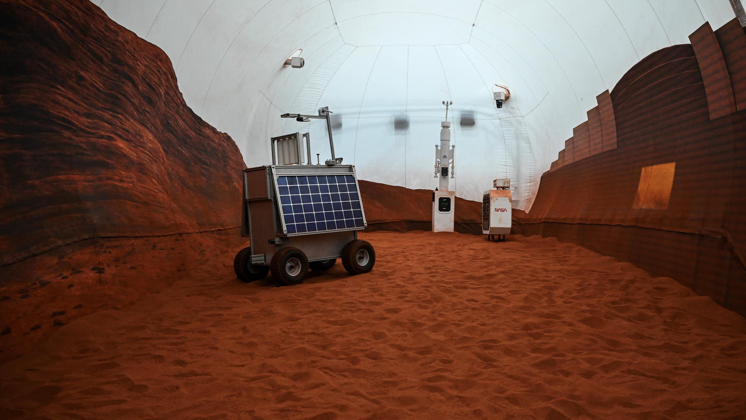 A rover with a solar panel on it sitting in a dome, with the floor covered in reddish dirt.