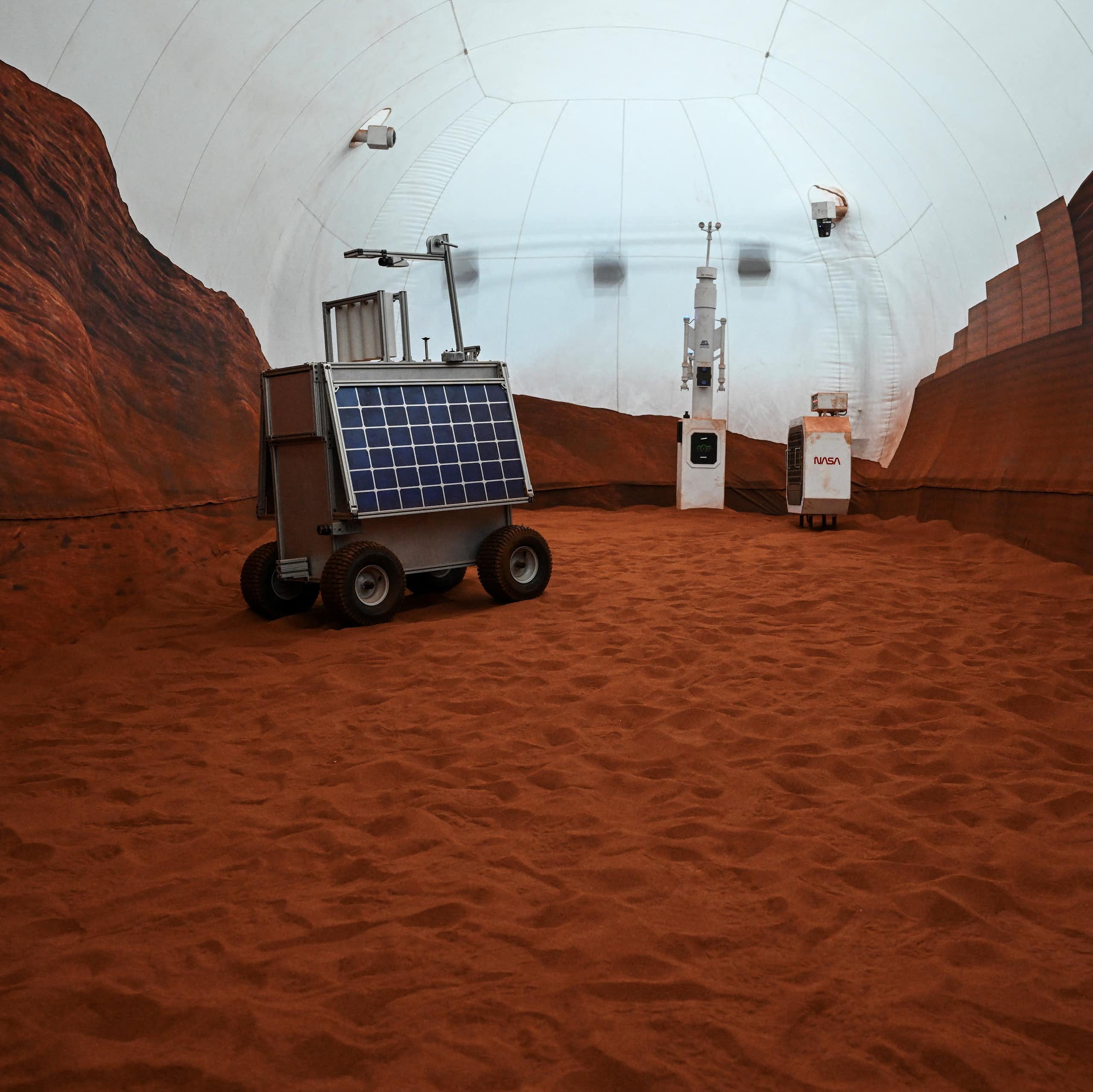 A rover with a solar panel on it sitting in a dome, with the floor covered in reddish dirt.