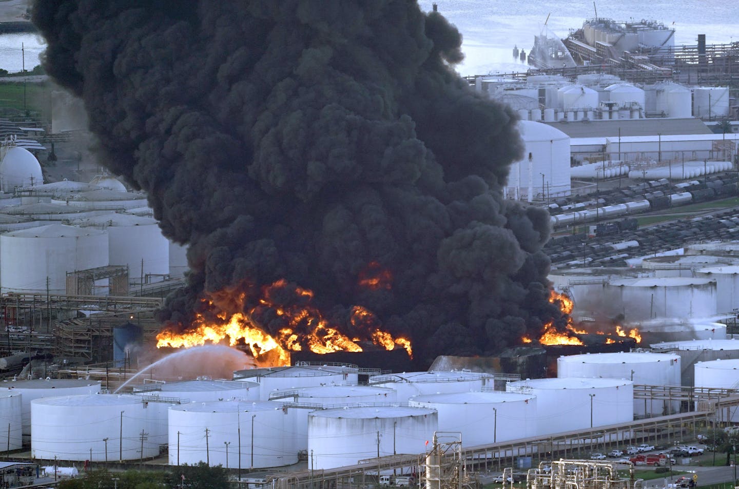 A fire at several large round buildings sends dark black smoke into the sky.