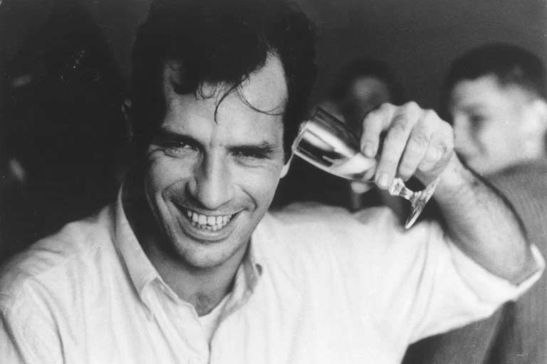 Jack Kerouac holding a champagne flute to his ear and smiling