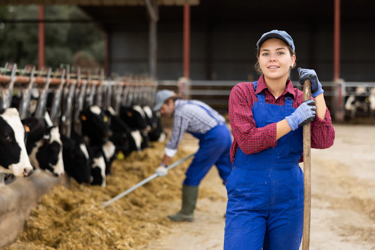 female farmer in blue dangarees smiling, cows in background in cowshed