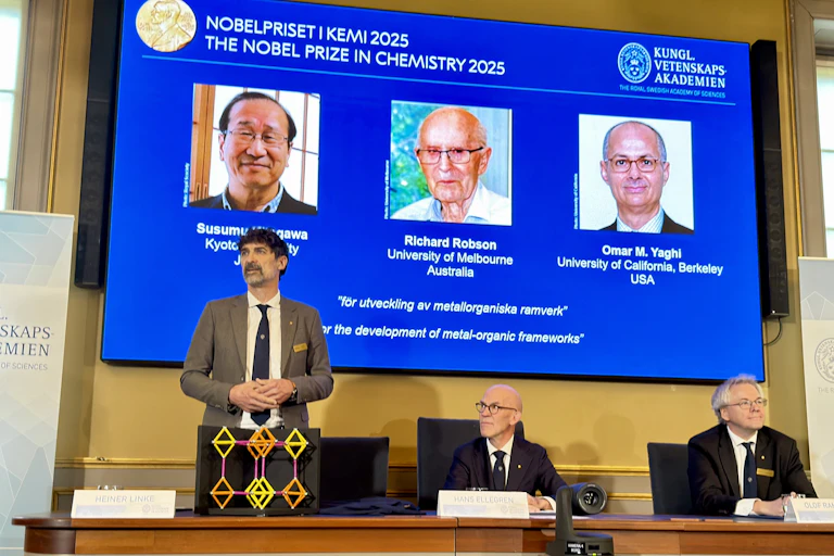 Two men sitting and one standing at a table in front of a screen with the headshots of the three Nobel Prize winners in chemistry in 2025 behind them. On the table sits a geometric model of a molecule.