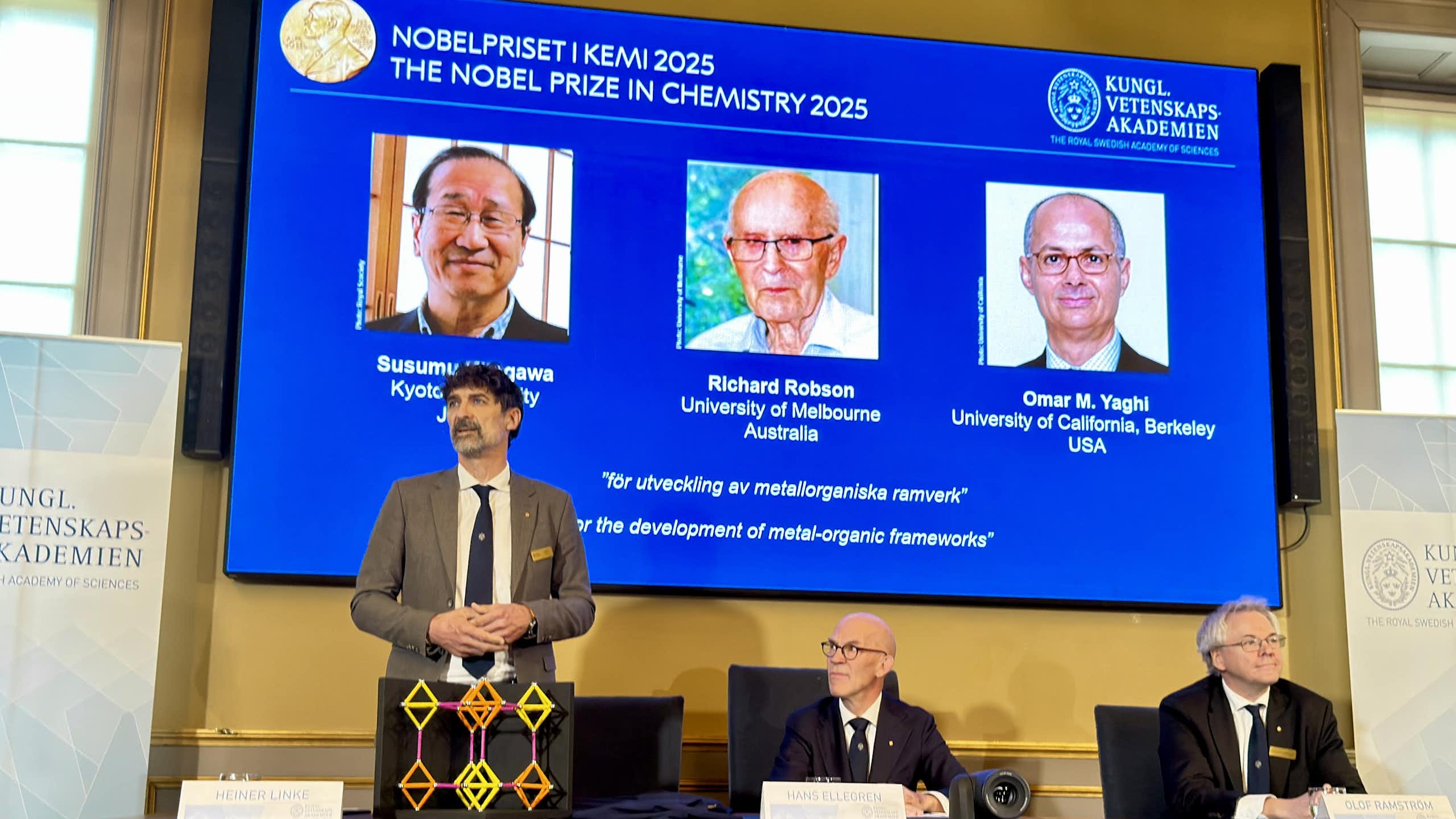 Two men sitting and one standing at a table in front of a screen with the headshots of the three Nobel Prize winners in chemistry in 2025 behind them. On the table sits a geometric model of a molecule.