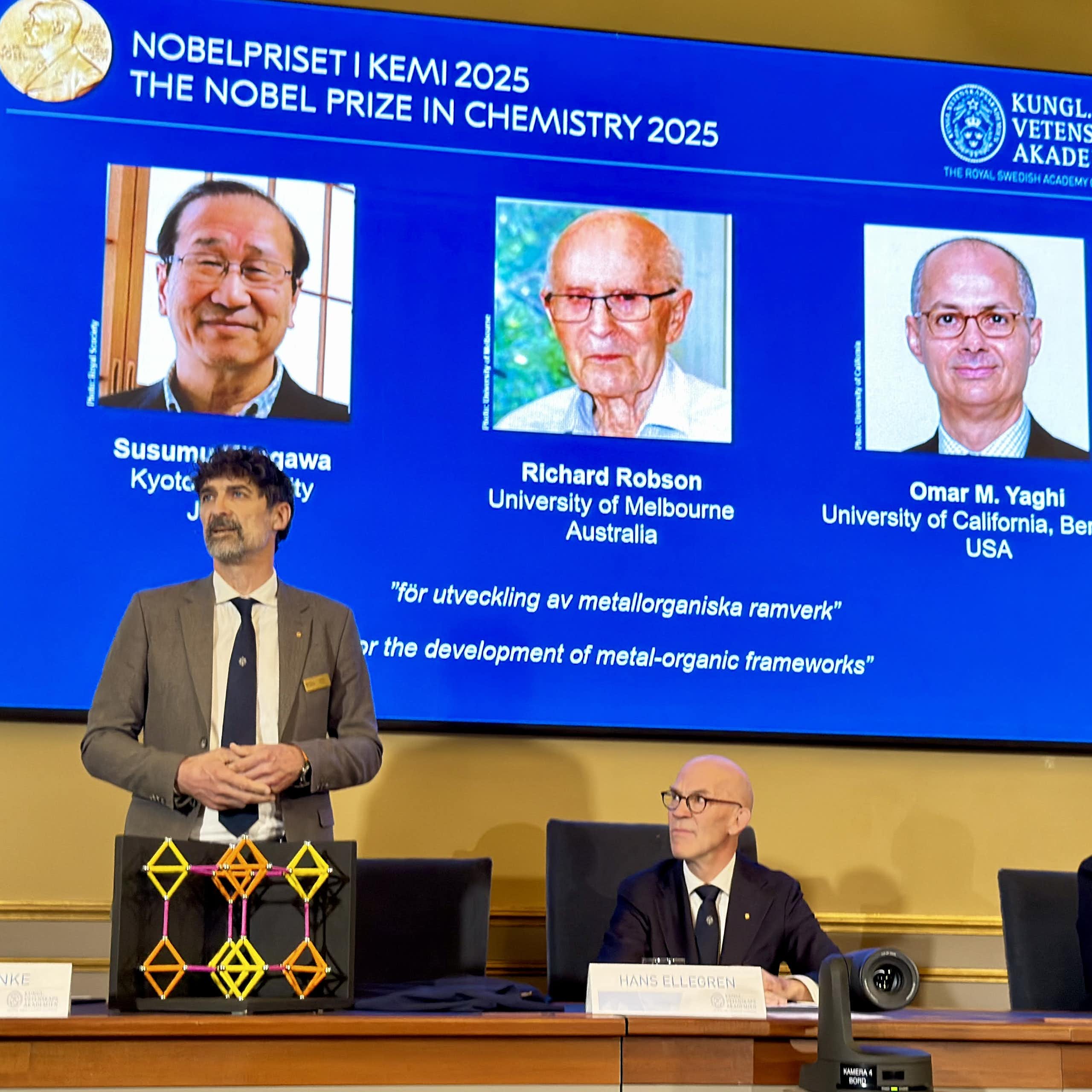 Two men sitting and one standing at a table in front of a screen with the headshots of the three Nobel Prize winners in chemistry in 2025 behind them. On the table sits a geometric model of a molecule.