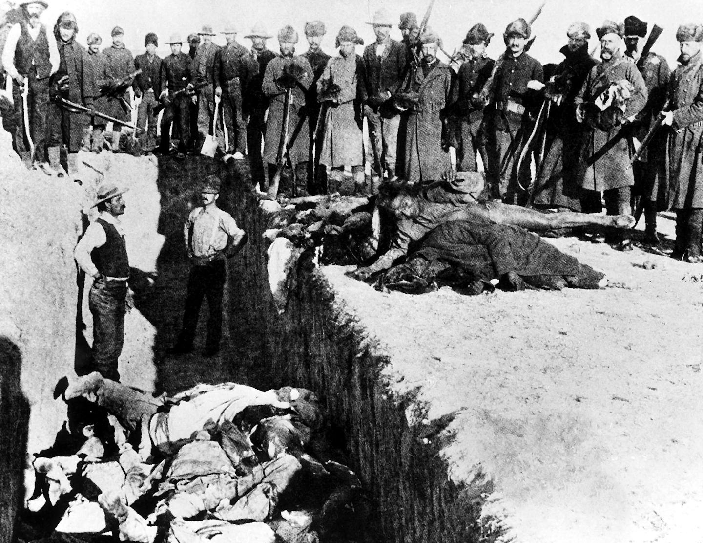 Black-and-white photo of soldiers standing around a ditch filled with corpses