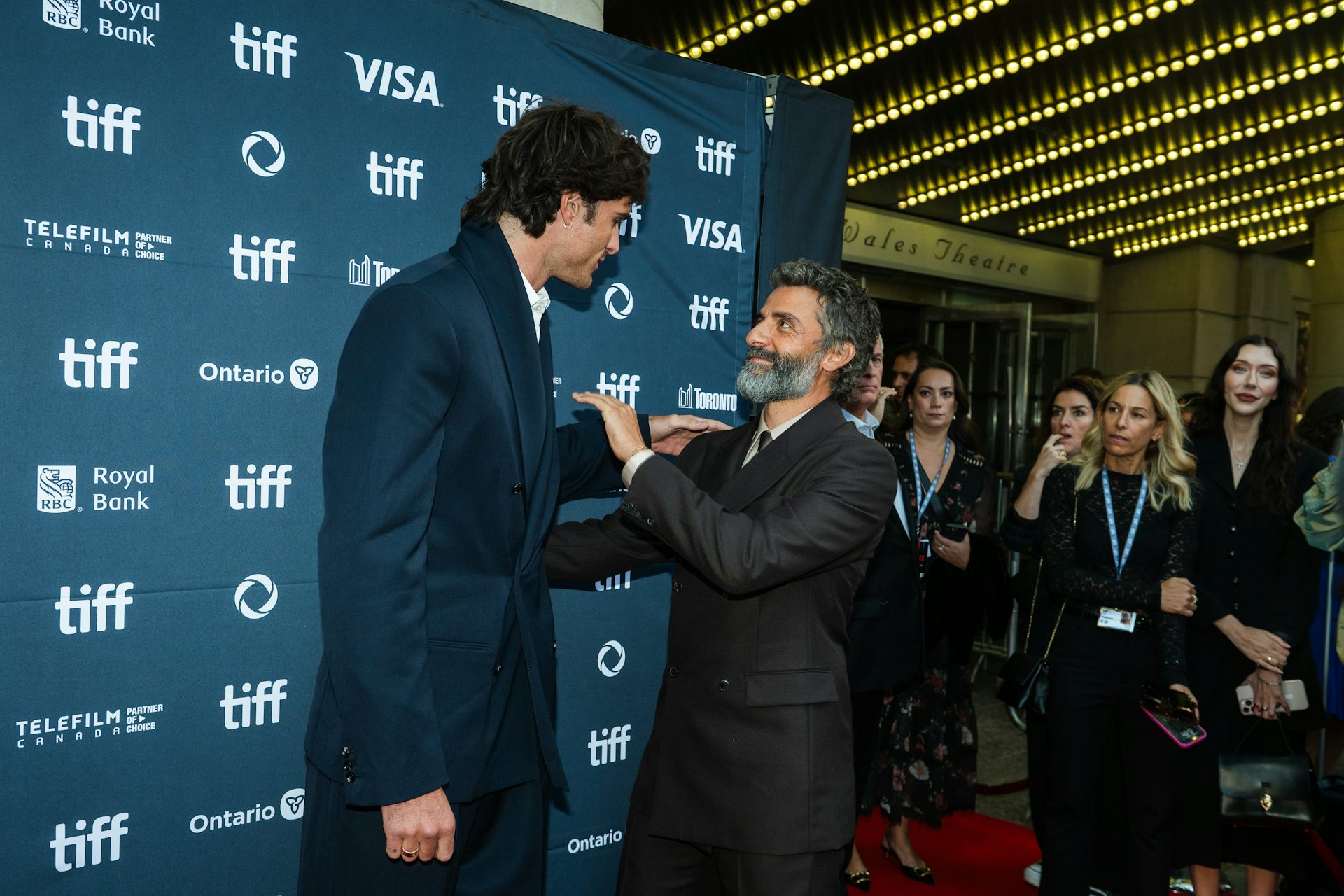 A very tall man bends towards a shorter man and they have their hands on one another's shoulders. 